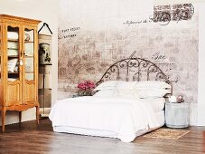 Bed with white linen and vintage metal frame against faded painting on wall and rustic, Baroque-style display cabinet