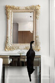 Black cat jumping down from mirrored glass console table below wall-mounted mirror with pale wooden frame carved with floral motifs