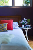 A double bed with a padded headboard under a bedroom window with decorative cushions and a cheerful bunch of flowers on a bedside table