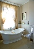 Freistehende Vintage Badewanne vor Fenster mit Vorhang und Designer Stuhl in schlichtem Bad