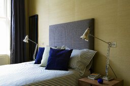 Bedroom with vintage lamps either side of bed with upholstered headboard and blue scatter cushions against sand-coloured fabric wallpaper