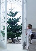 Blick auf Weihnachtsbaum durch offen stehende Flügeltür und Kind auf Sofa