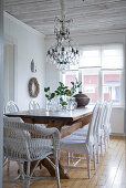 Rustic dining area with white chairs around solid wooden table below crystal chandelier