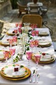 A table laid for a celebration meal outdoors, with coloured wine glasses and plates with a flower design on a white tablecloth