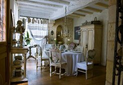 Festively decorated table with floor-length tablecloth, collection of antique chairs and chandelier in austerely restored country house