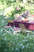 Voyeuristischer Blick durch das verschwommene Gartengrün auf verschiedene Stühle um einen sommerlich gedeckten Tisch mit Bougainvillea-Topf