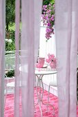 Blick durch die Bahnen eines pastellvioletten Vorhangs auf Terrassen-Sitzecke im femininen Shabbylook mit zierlichen Bistromöbeln und Deko in Rosé- und Pinktönen