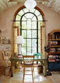 Old wooden table with a glass top in front of a large arched window in the attic with wooden beam ceiling