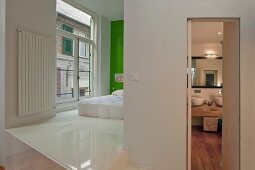 Offener Schlafbereich mit weißem Kunstharzboden und Bett vor grün getönter Wand neben Bad mit offener Tür und Blick auf Waschtisch