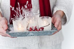 Hands holding tray of lit tealights in ornamented holders and holly berries