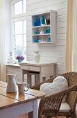 View across kitchen table and wicker chair of console table and small shelving unit on white wooden wall