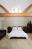 Weiß bezogenes Doppelbett und braune Holzkommoden in ausgebautem Dachraum mit offener Holzbalkenkonstruktion