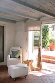 Pale armchair and ottoman on castors in front of open terrace door in living room with Mediterranean terracotta tiles and exposed ribbed ceiling