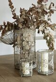 Getrocknete Blätterzweige in ehemaligem Vogelkäfig in Vintage Stil und Windlichter mit weissen Kerzen
