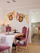 Chairs with pink upholstery at dining table in corner of room with coats of arms on wall next to doorway leading to kitchen