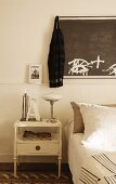 Antique bedside table with retro-style table lamp and dark artwork on wall above bed