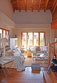 View past staircase into lounge area with pale sofa set in front of balcony window in open-plan interior with wooden ceiling