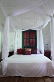 Airy, white fabric over canopied bed in front of green-painted window with closed shutters