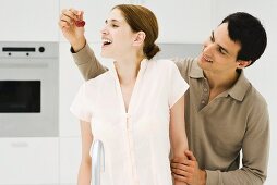 Man feeding woman cherry, both smiling, looking away