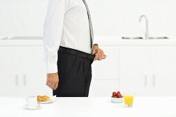 Man standing in kitchen beside two breakfast options, holding waistline of pants, cropped