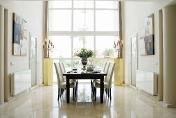 Dark wood table and chairs with white upholstery in front of floor-to-ceiling window in elegant setting