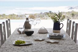 Für einfache Mahlzeit gedeckter Tisch mit Korbflasche und Krugvase; im Hintergrund Blick auf Meeresküste