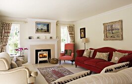 Traditional furniture with upholstery in different plain colours in bright interior with fireplace and landscape painting; gathered curtains and rug in centre of room.