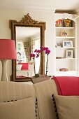 Mirror with antique-style gilt frame behind beige sofa; lampshade and folded blanket in a rich pink; feminine atmosphere in living area