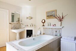 Dreiseitig zugängliche Badewanne mit weiss lackierter Holzverkleidung und Säulenwaschbecken neben kleinem Kamin