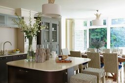 Offene Küche im eleganten maritimen Landhausstil mit abgerundetem Küchenblock und Korbstühlen an gedeckter Tafel im Hintergrund