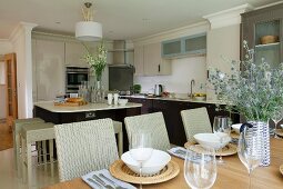 Elegant kitchen with wicker stools at kitchen island and festively set table with basketwork place mats in foreground