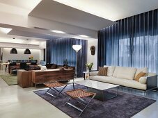 Loungebereich mit eleganten Hockern und weißem Sofa vor Fenster mit geschlossenem Vorhang in offenem Wohnzimmer