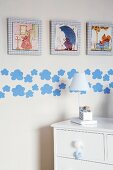 Detail of child's bedroom - table lamp on white chest of drawers against wall with framed pictures and applied pattern of clouds