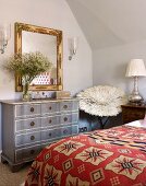 Bed with ethnic bedspread and pale grey vintage chest of drawers in attic bedroom