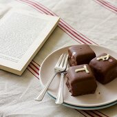 Petit Fours auf Teller neben Kuchengabeln und aufgeschlagenes Buch auf Leinentischdecke