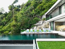 Holzdeck mit Liegestühlen am Pool eines in den Fels gebauten, zeitgenössischen Wohnhauses; Blick auf bewaldeten Hang