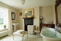 Bathroom with open fireplace and white tiled floor, large mirror above full bathtub, towels on ottomans and washstand in background