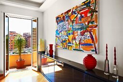 Abstract painting above glossy black sideboard and open balcony door with view of city