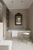 Framed mirror over a bathtub in a bathroom with mosaic tiles; green chair with bath towel