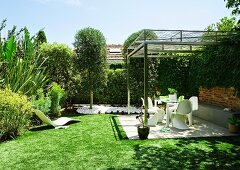 Garden with paved terrace and light pergola; tall hedges and various garden plants in background