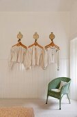 Antique Hungarian linen smocks and shirts hung decoratively on a white wall above a green Lloyd Loom chair