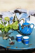 A blue porcelain teapot with silver decoration and matching tea tumblers on an old, round, metal garden table