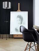 Antike Staffelei mit darauf stehendem, modernen Portrait vor schwarzer Wand; im Vordergrund schwarze Charles Eames Stühle