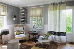 Einfaches Wohnzimmer mit hellgrauen Wänden und weisser Wandverkleidung; in der Ecke ein Lesessel mit weichen Kissen und ein antiker Beistelltisch