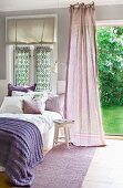 Bequemes Bett mit lila Bettdecke neben wehendem, mauvefarbenen Vorhang vor offener Gartentür