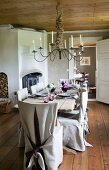 Rustic dining table and chairs with loose covers of rough linen in pleasant room with pale wooden ceiling and lit candles in large chandelier