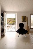 Klassikersessel mit schwarzem Polster vor offener Terrassentür in schlichtem Wohnraum mit weisser Holzdecke