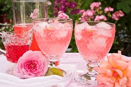 Rosensirup mit Eiswürfeln in zwei Gläsern und Rosenblüten auf einem Tisch im Freien
