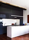 Tall fitted kitchen with glossy, black doors and white counter below suspended strip light
