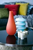 Artistic, coloured glass vases and small, finely ornamented beakers on dark, steel and glass table; scatter cushions in blurred background